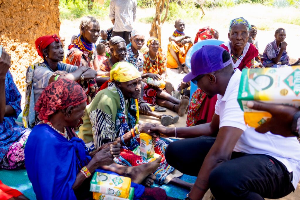 Elders seated in a circle outdoors, receiving packs of food from the team